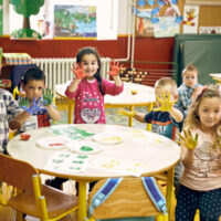 A group of kids, some of them sitting on the chairs and the remaining kids just standing around the round table, each showing their hands filled with colors.
