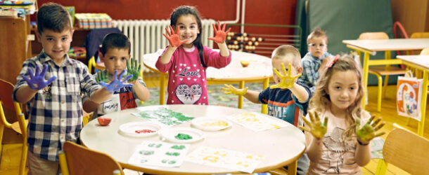 A group of kids, some of them sitting on the chairs and the remaining kids just standing around the round table, each showing their hands filled with colors.