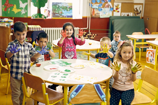 A group of kids, some of them sitting on the chairs and the remaining kids just standing around the round table, each showing their hands filled with colors.